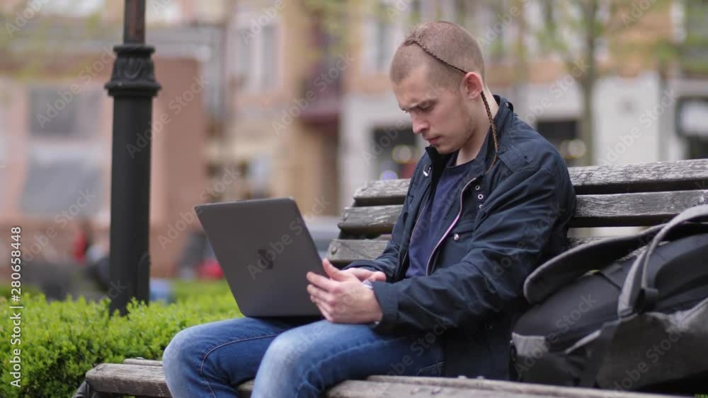 Serious young man using laptop pc to study online sitting on bench in city park. Handsome focused male student preparing for college classes remotely outdoor surfing Internet on laptop computer Serious young man using laptop pc to study online sitting on bench in city park. Handsome focused male student preparing for college classes remotely outdoor surfing Internet on laptop computer