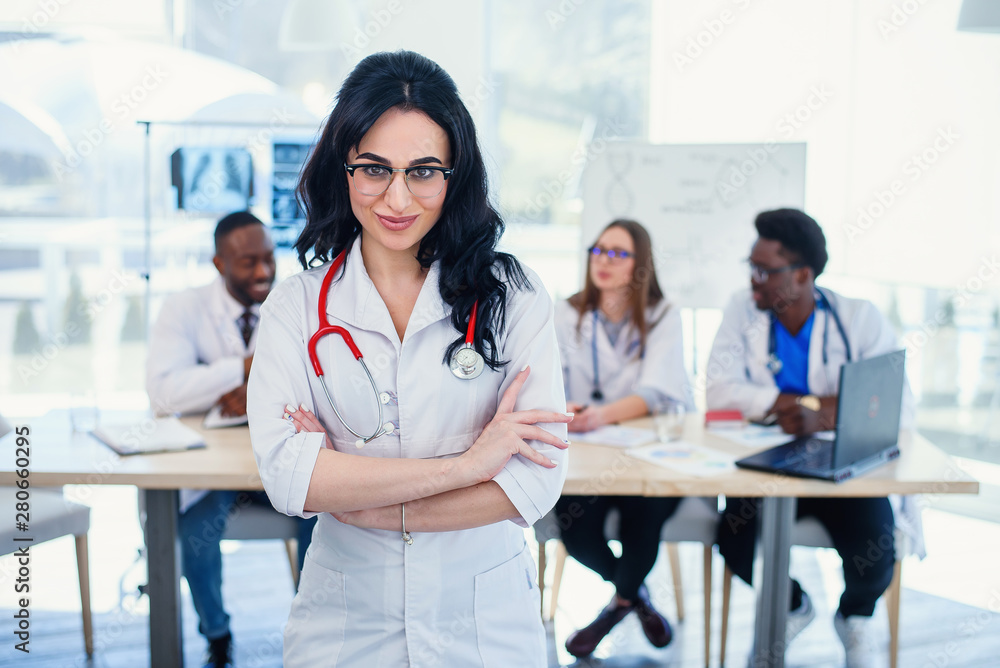 Fototapeta premium Smiling medical doctor woman with stethoscope standing in front of medic team at hospital. Attractive young female doctor in white coat and glasses looking at the camera.
