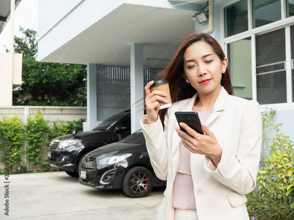 Fototapeta premium Asian woman holding coffee cup and using smartphone while standing in front of house. Portrait of smiling asian businesswoman