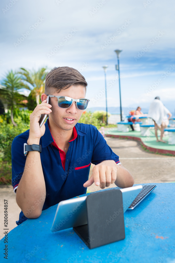 Young student speaks on the phone while preparing his work for the university. He is working outdoors in a park with a tablet, smartwatch  and a smartphone.