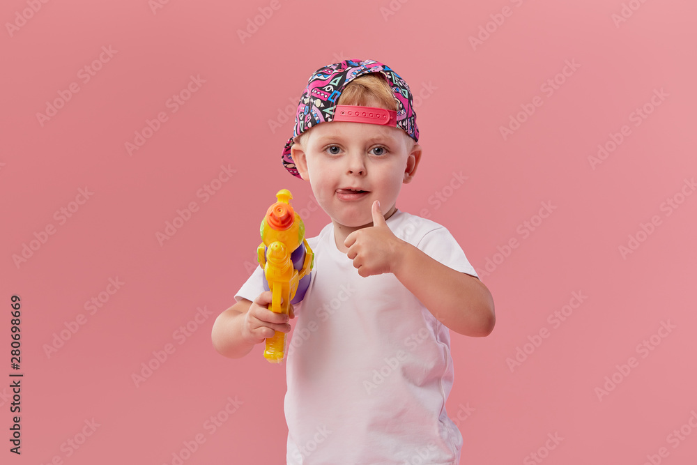 Happy little toddler boy in summer clothes holds toy water gun isolated on pink wall background. Children studio portrait. People childhood lifestyle concept. Happy little toddler boy in summer clothes holds toy water gun isolated on pink wall background. Children studio portrait. People childhood lifestyle concept.