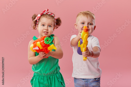 Happy little toddler boy and girl in summer clothes holds toy water guns isolated on pink wall background. Children studio portrait. People childhood lifestyle concept. 