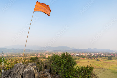 Flag fluttering atop the Pha Poak hill. Beautiful view of fields, Vang Vieng town and mountains from above in Laos, on a sunny day. Copy space.