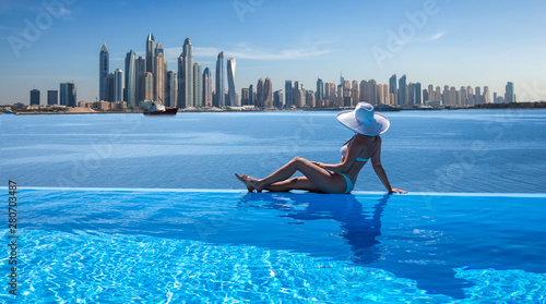 Photography Beautiful panorama of Dubai Marina skyline in a background with a pool, deck chair and woman with a white hat