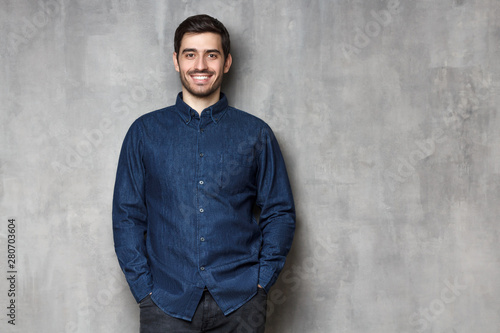 Young man smiling, dressed in blue denim shirt, standing isolated on gray wall background with copy space