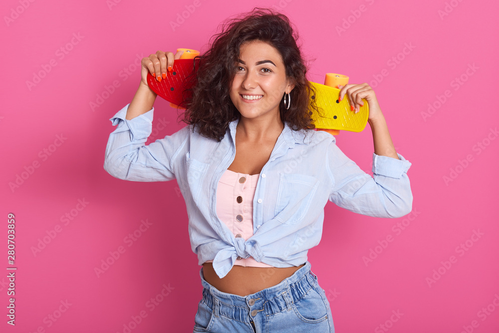 Close up of brunette charming woman with tender smile, holding skateboard on her shoulders, wearing t shirt, shirt and jeans, enjoys hobby, isolated over pink background, ready to skate in park. Close up of brunette charming woman with tender smile, holding skateboard on her shoulders, wearing t shirt, shirt and jeans, enjoys hobby, isolated over pink background, ready to skate in park.