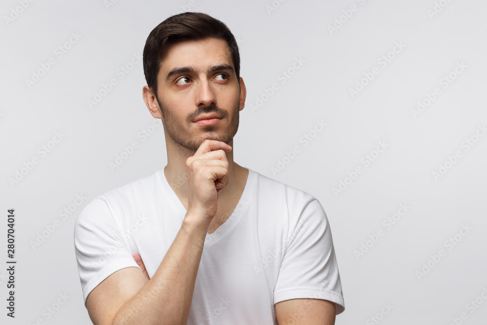 Young guy isolated on grey background standing in blank T-shirt against wall, pressing fist to chin as if dreaming of something, looking pensive and happy Young guy isolated on grey background standing in blank T-shirt against wall, pressing fist to chin as if dreaming of something, looking pensive and happy