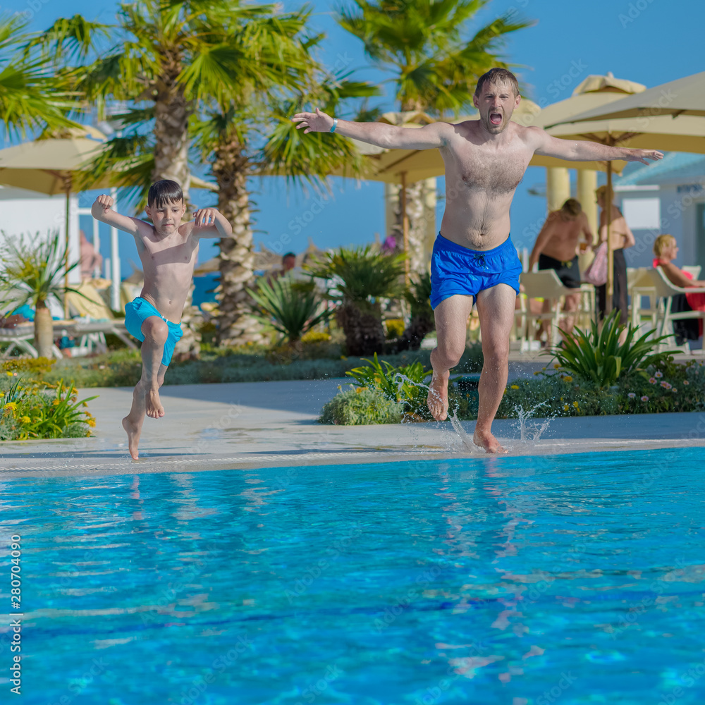Cute European boy and his dad having fun on hotel pool on summer holidays. They are jumping in to the pool with open wide hands.