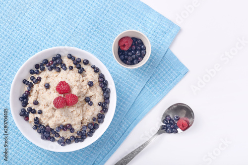 Photo of deep plate of oatmeal with strawberries and raspberries stand on delicate white wooden background on which lies blue cotton towel. Healthy eating, breakfast, healthy food and diet concept.