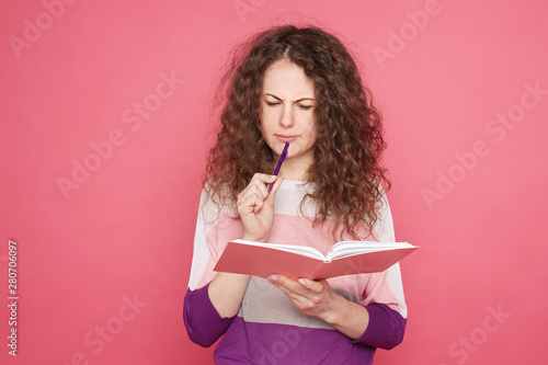 Fashionable beautiful talented college girl focusing on home assignment, writing essay in pink notebook, reading learning new words, has concentrated face expression, posing against pink studio wall.