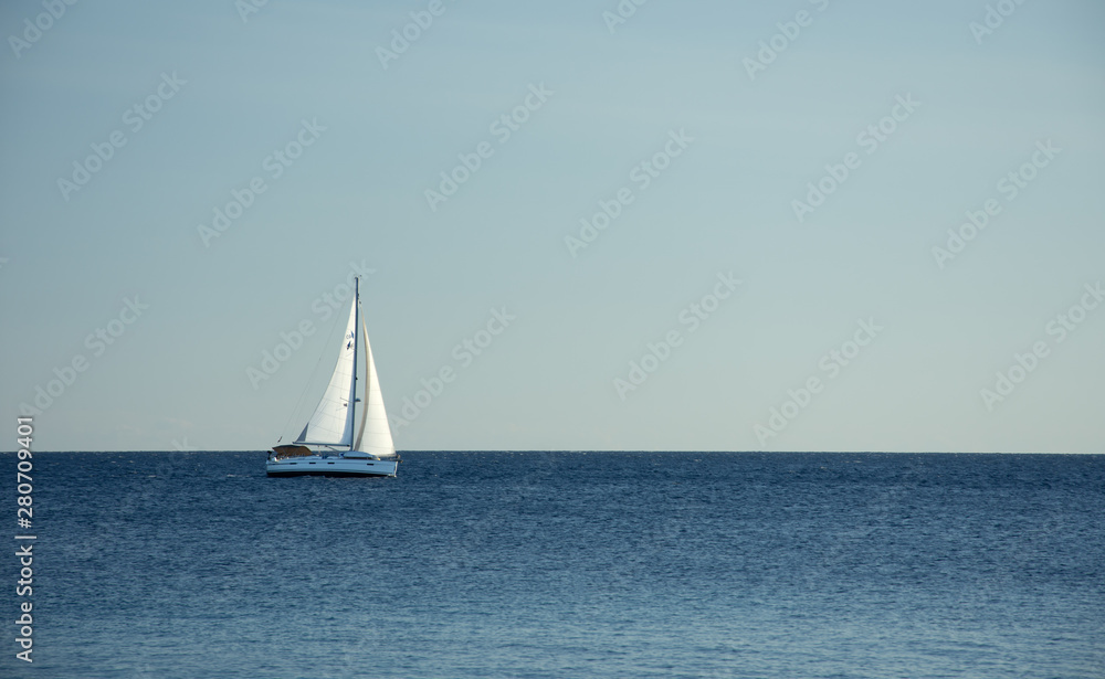 Summer day on the Adriatic sea.  In the distance, a white sailboat is sailing along the sea in the windy and sunny day. Horizontal, minimalism photo. In shades with blue. With a lot of space for text.