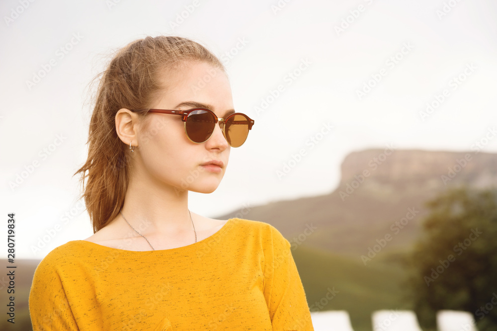 Portrait of a young attractive serious girl in sunglasses against the background of rocks and sky. Seriousness in the millenial generation Portrait of a young attractive serious girl in sunglasses against the background of rocks and sky. Seriousness in the millenial generation