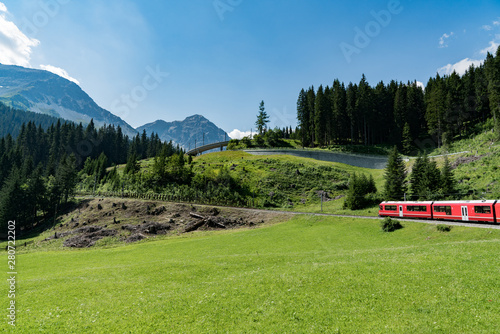 red Rhaetian railway train in a green summer mountain landscape in the Swiss Alps