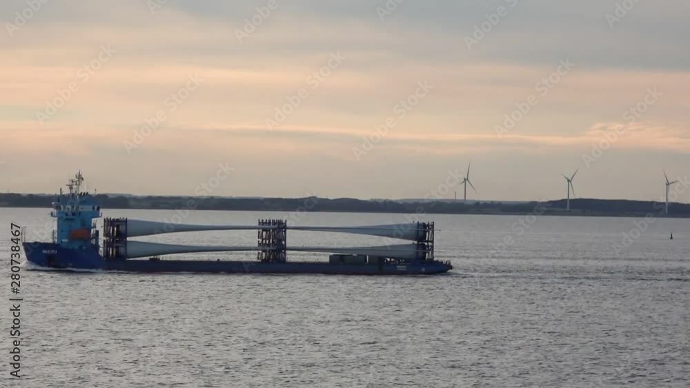 RIGA/LATVIA - JUNE 21 2019: Long hopper barge sails on sea surface against land covered with high windmills row under pink sky at sunset on June 21 in Riga RIGA/LATVIA - JUNE 21 2019: Long hopper barge sails on sea surface against land covered with high windmills row under pink sky at sunset on June 21 in Riga