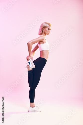 Fototapeta Naklejka Na Ścianę i Meble -  Strong and flexible. Back view of sporty and cute slim woman in sportswear stretching her legs while standing against pink background in studio