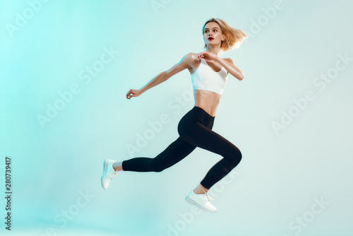 No limits! Sporty and young woman with perfect body in white top and black leggings jumping against blue background in studio