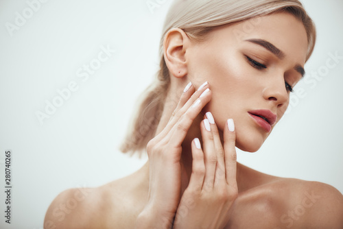 Perfect skin. Portrait of gorgeous blonde woman touching her soft glowing skin while standing against grey background
