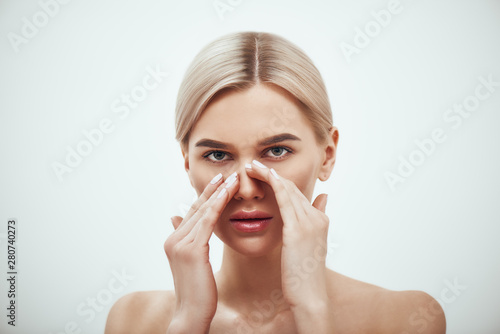Rhinoplasty - nose surgery. Portrait of attractive blonde woman touching her nose and looking at camera while standing against grey background photo