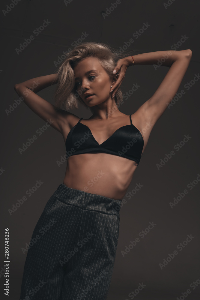 Seductive portrait of young sexy girl with blonde hair in lingerie and trousers on black background in room Seductive portrait of young sexy girl with blonde hair in lingerie and trousers on black background in room