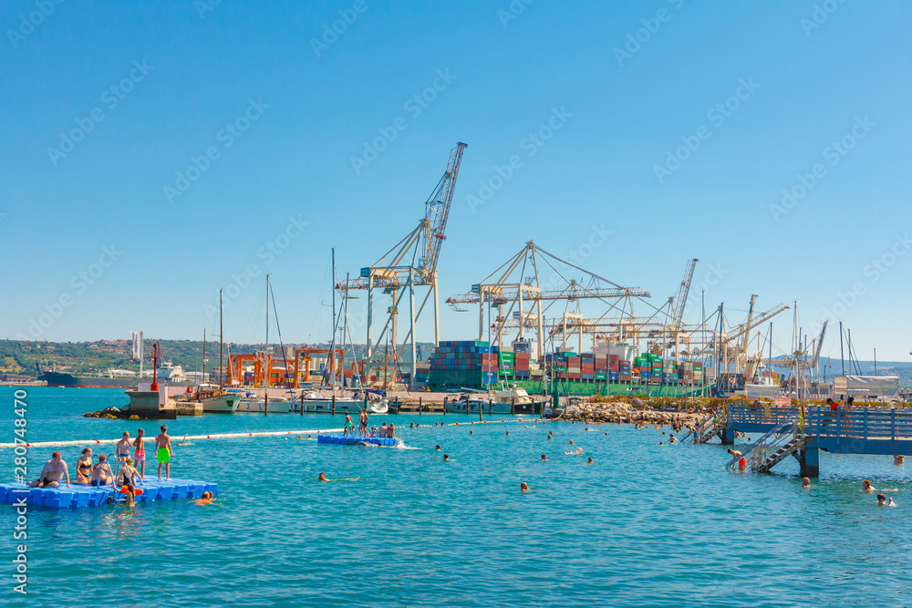 People swimming on the beach in front of harbor cranes and luxury cruise liner. Koper, Slovenia