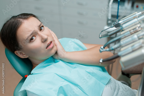 Patient in dental chair. Beautiful young woman having dental treatment at dentist's office. Girl touch her cheek suffering from painful toothache.