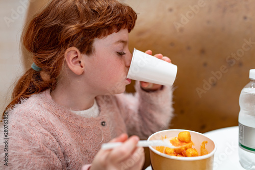 Bambina capelli rossi, che mangia e beve di gusto in una ristorazione, bar, fast food photo
