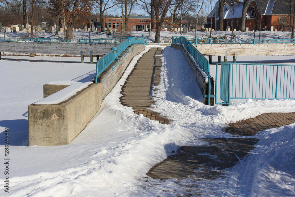Obraz premium Snow & ice covered bridge with turquoise fencing, crossing a snow covered frozen lake. Trees and brick buildings in the backgroudn.