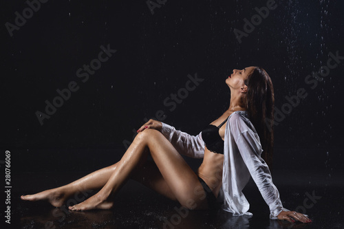 attractive beautiful athletic girl model in black underwear and white shirt sitting on floor under water droplets on black background