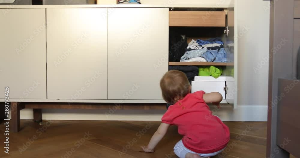 Back view of little baby opening cabinet doors and looking inside while sitting on floor in room Back view of little baby opening cabinet doors and looking inside while sitting on floor in room
