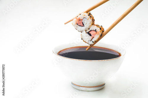 Sushi held in chopsticks over a bowl with sauce on a white background