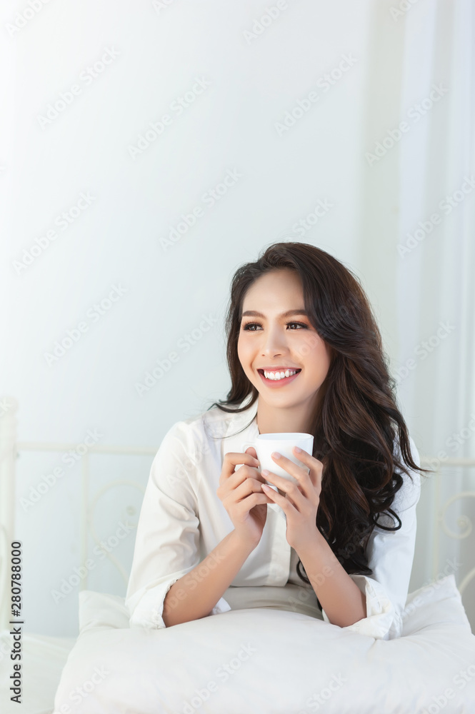 A beautiful asian woman in a white shirt and coffee cup that is smiling happily in the white bed. A beautiful asian woman in a white shirt and coffee cup that is smiling happily in the white bed.