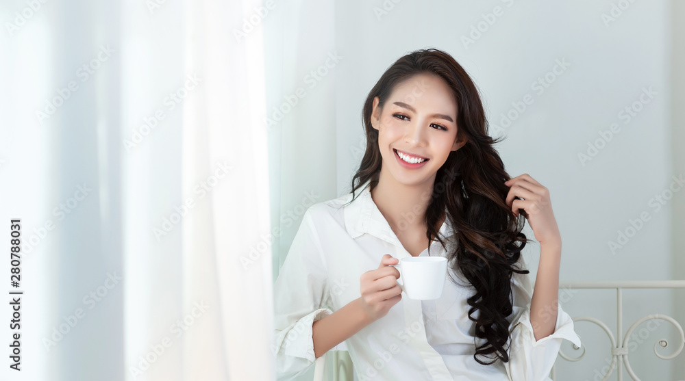 A beautiful asian woman in a white shirt and coffee cup that is smiling happily in the white bed.