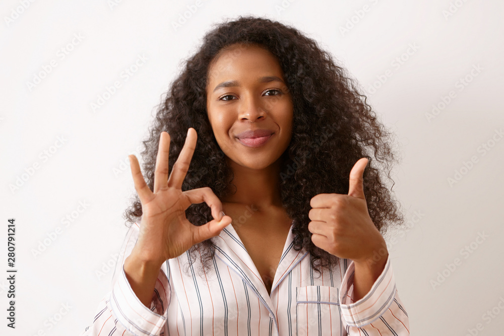 Joy, positiveness and body language. Beautiful happy young girl with curly black hair posing isolated dressed in silk pajamas, making thumbs up gesture and showing ok sign, smiling Joy, positiveness and body language. Beautiful happy young girl with curly black hair posing isolated dressed in silk pajamas, making thumbs up gesture and showing ok sign, smiling