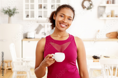 Portrait of cute African American girl with gathered hair posing in kitchen interior with cup of tea. Attractive happy dark skinned female drinking coffee, looking at camera with toothy smile photo