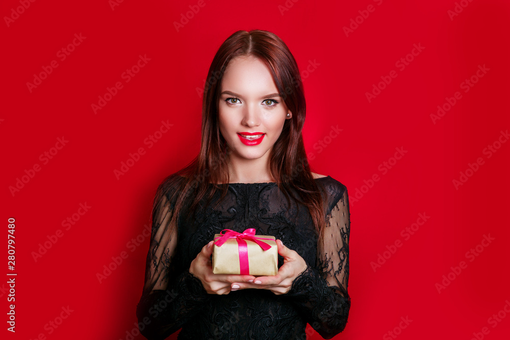 Happy woman in black dress holding gift box and looking at the camera while enjoys over red background Happy woman in black dress holding gift box and looking at the camera while enjoys over red background