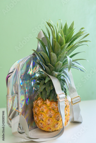 Pineapple with holographic bag on white wooden table over neo mint background. Tropical summer vacation, travel, back to school and beach party. photo