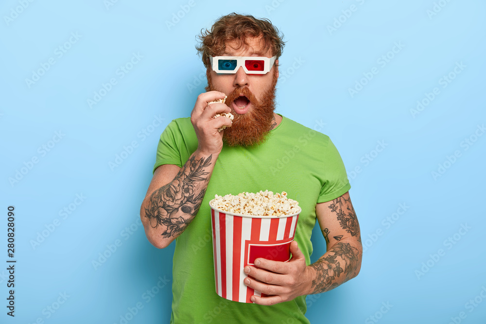 Φωτογραφία Isolated shot of handsome man has tattoo, ginger hair, watches movie, involved w