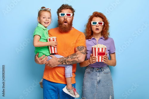 Friendly family of mum, dad and child visit film festival in cinema, watch psychological thriller or horror film, wear 3d glasses, eats fast food from bucket. Redhead three spectators enjoy movie photo