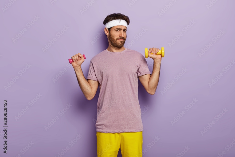 Young man looks unpleasantly at camera, raises two dumbbells, has fitness exercise, trains muscles on hands, has sporty body, isolated over purple wall in gym. Workout with weights. Healthy lifestyle Young man looks unpleasantly at camera, raises two dumbbells, has fitness exercise, trains muscles on hands, has sporty body, isolated over purple wall in gym. Workout with weights. Healthy lifestyle