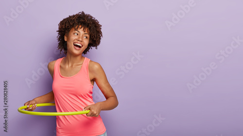 Foto Sportive carefree woman with curly hairstyle, spins yellow plastic hoop, has gym