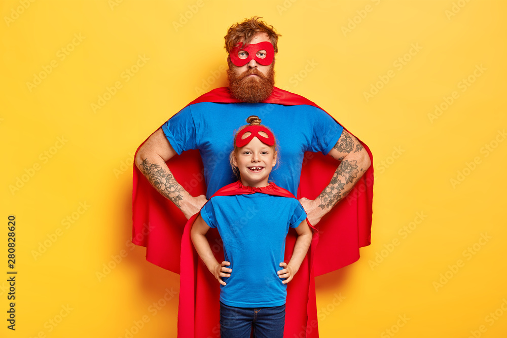 Photo of confident ginger father and daughter keep hands on waists, pretend being superhero and defend people, wear special costumes, isolated on yellow background. Family team ready to work Photo of confident ginger father and daughter keep hands on waists, pretend being superhero and defend people, wear special costumes, isolated on yellow background. Family team ready to work