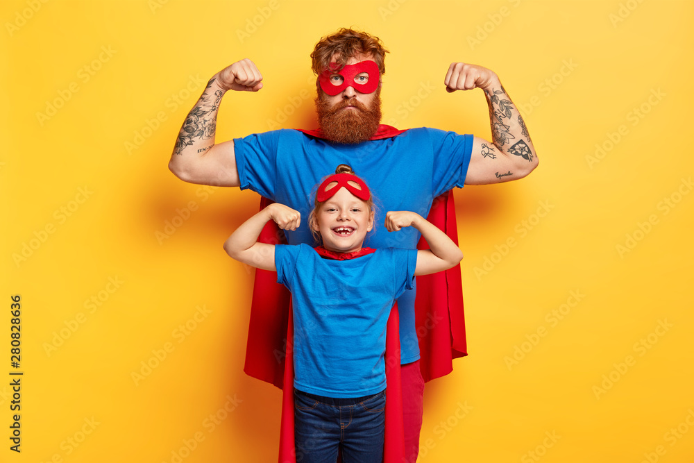 Superhero family. Strong father and small child show biceps, pretend being superman, wear red masks, blue t shirts and cloaks, motivated for successful future, stand agaisnt yellow studio wall - obrazy, fototapety, plakaty 