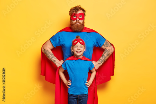 Photo of confident ginger father and daughter keep hands on waists, pretend being superhero and defend people, wear special costumes, isolated on yellow background. Family team ready to work