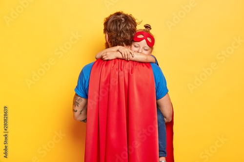 Horizontal shot of caring father carries small daughter, receives warm hug, wear red cloak and mask, play superhero game, dressed in special costumes, isolated on yellow wall. Male defender with kid