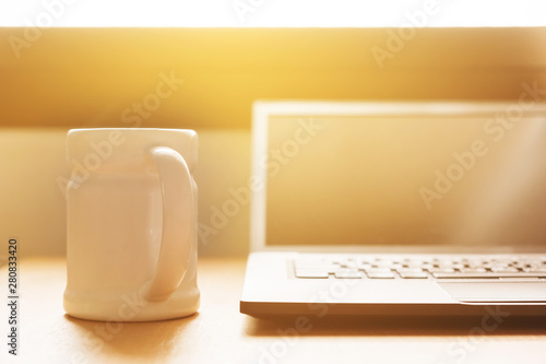 laptop is on the desk at home near window in the morning. there is a cup of coffee for refresh you from awake. the sun is shining from outside onto the desk.