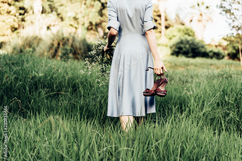 Rear view of a woman in a dress walking along a meadow barefoot with shoes in her hand