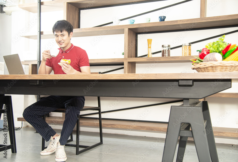 Asian smiling young man with casual red t-shirt enjoy having breakfast, eating sandwich, Young man cooking food and drink in the loft style kitchen room Asian smiling young man with casual red t-shirt enjoy having breakfast, eating sandwich, Young man cooking food and drink in the loft style kitchen room