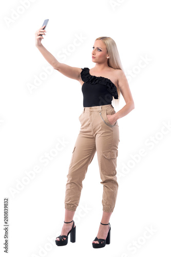 Confident serious young blonde woman in sleeveless black top taking selfies. Full body isolated on white background. 