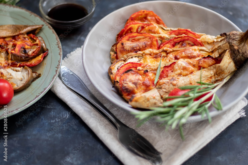 Dietary fan of eggplant white filled with tomatoes and cheese. Nearby are cherry tomatoes, rosemary branches. It is worth soy sauce and metal appliances. On a dark background. Dietary fan of eggplant white filled with tomatoes and cheese. Nearby are cherry tomatoes, rosemary branches. It is worth soy sauce and metal appliances. On a dark background.
