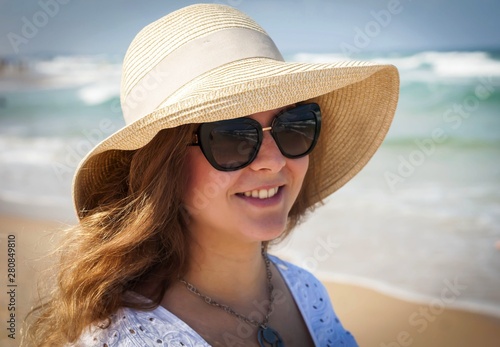 Happy beautiful young Caucasian Russian woman in her late 20s in a straw hat with sunglasses on a hot summer day at the seaside with sea waves on her background. Summer vacation at the sea concept